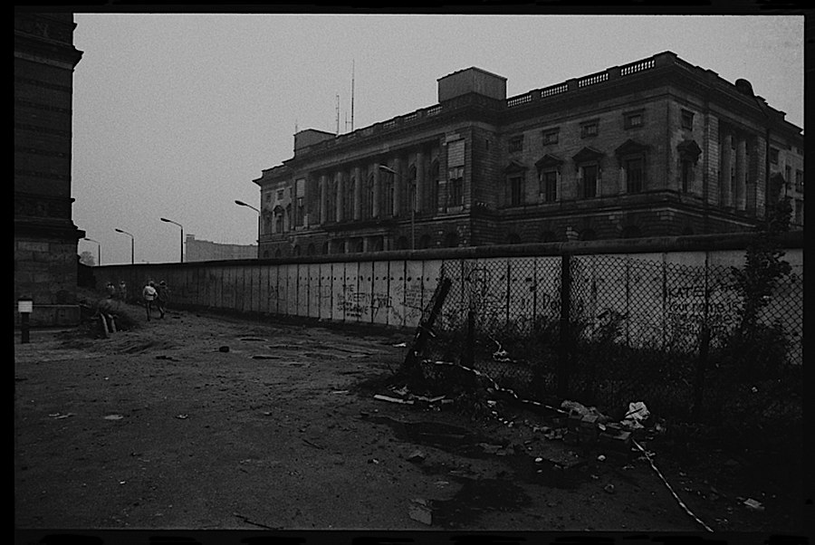 Photographie noir et blanc d'un batiment quelque peu caché par le mur ainsi que d'enfants semblant jouer à côté du-dit mur