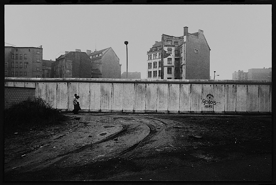Photographie noir et blanc quelque peu éloignée d'une partie du mur laissant apparaitre une maison de deux ou trois étages et de deux personnes marchant à côté du mur</a>
